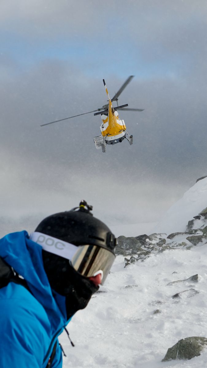 A rescue helicopter hovers above a snowy landscape, while a person in a blue jacket and helmet observes from the foreground. The scene is set in a mountainous, winter environment with overcast skies.