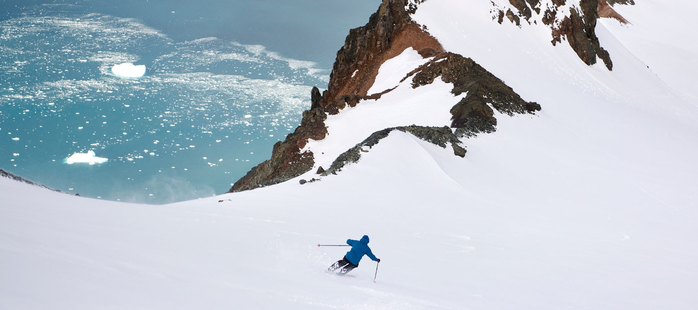A skier in a blue jacket descends a snowy slope, with a glacial blue lake and rocky cliffs visible in the background.