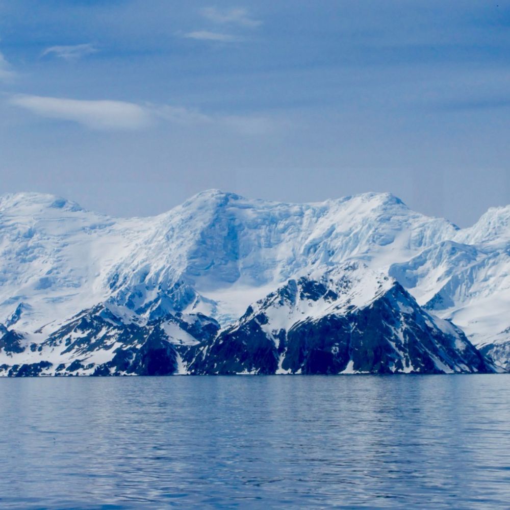 Snow-covered mountains rise dramatically under a clear blue sky, reflecting on a calm, icy sea. The landscape showcases the stark beauty of a polar region.