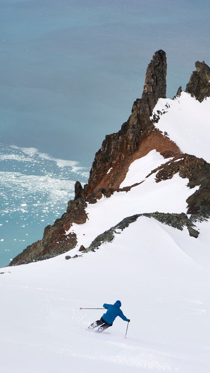 A skier in a blue jacket glides down a snowy slope, with a rocky outcrop rising sharply nearby. Below, turquoise waters reflect the light.