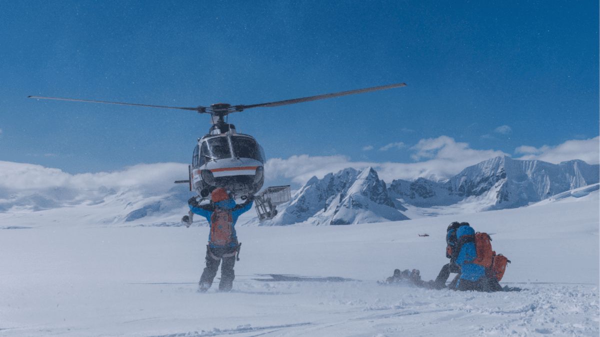 Helicopter landing on a snowy plain in a mountain range as people in winter gear wait on the ground.