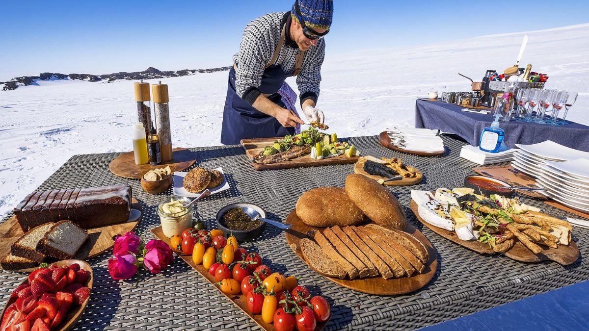 A chef prepares an elaborate outdoor feast on a table covered with a variety of gourmet dishes, including fresh vegetables, artisan breads, and desserts, against a stunning snowy landscape.