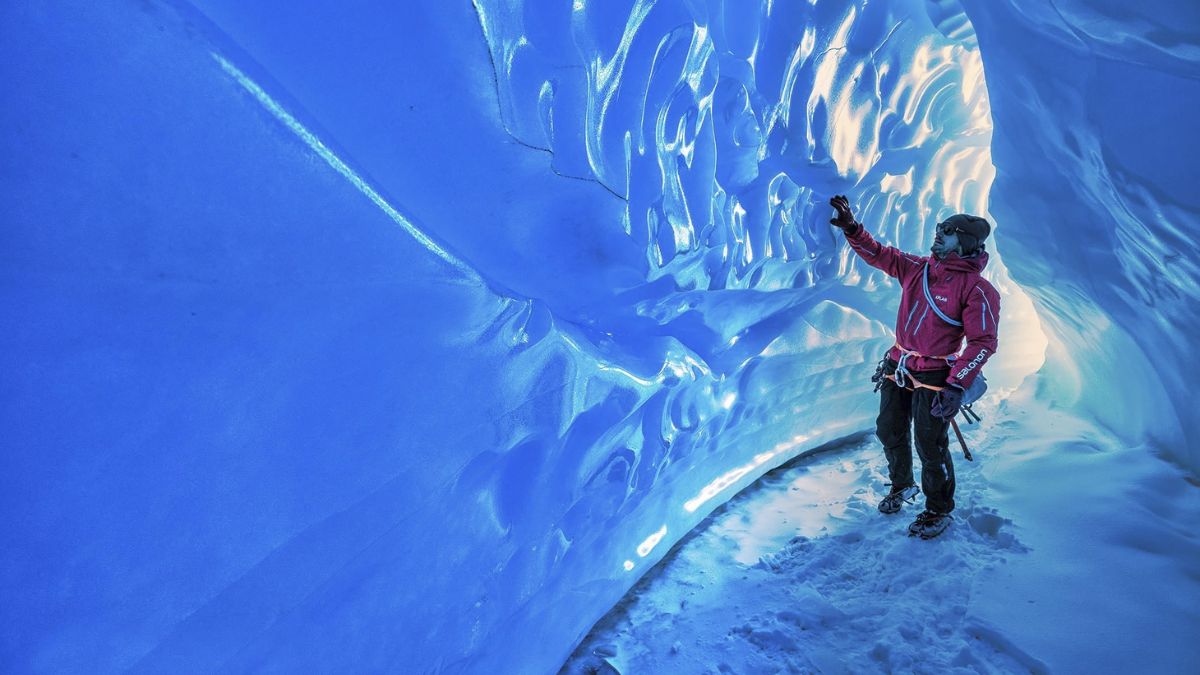 A climber in a red jacket explores an icy cave, illuminated by blue light reflecting off the crystalline ice formations. Snow covers the ground, and the climber reaches out to touch the textured ice wall.