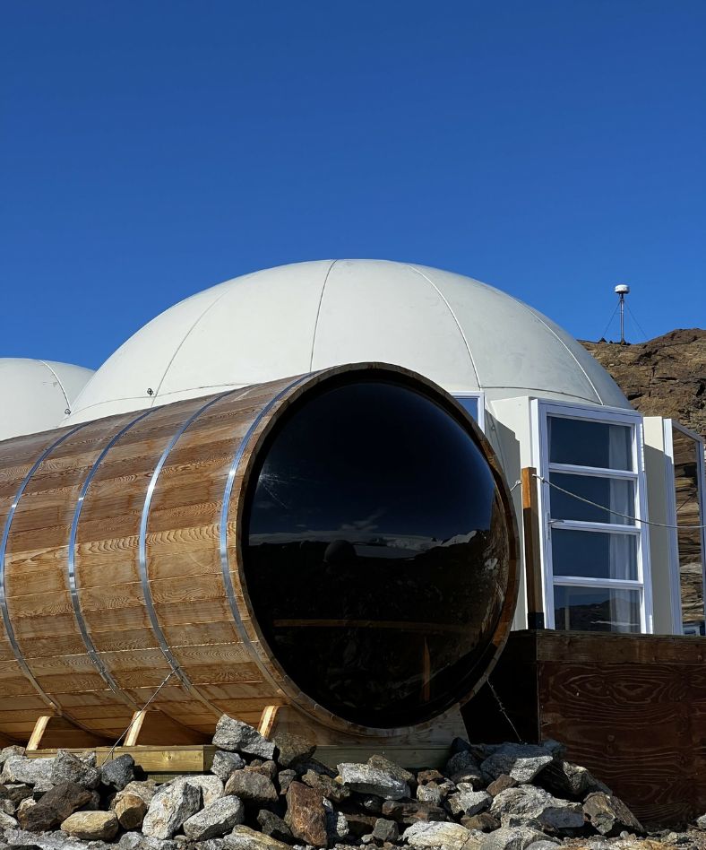 A modern geodesic dome structure accompanied by a large wooden cylinder set against a clear blue sky, with rocks visible in the foreground.