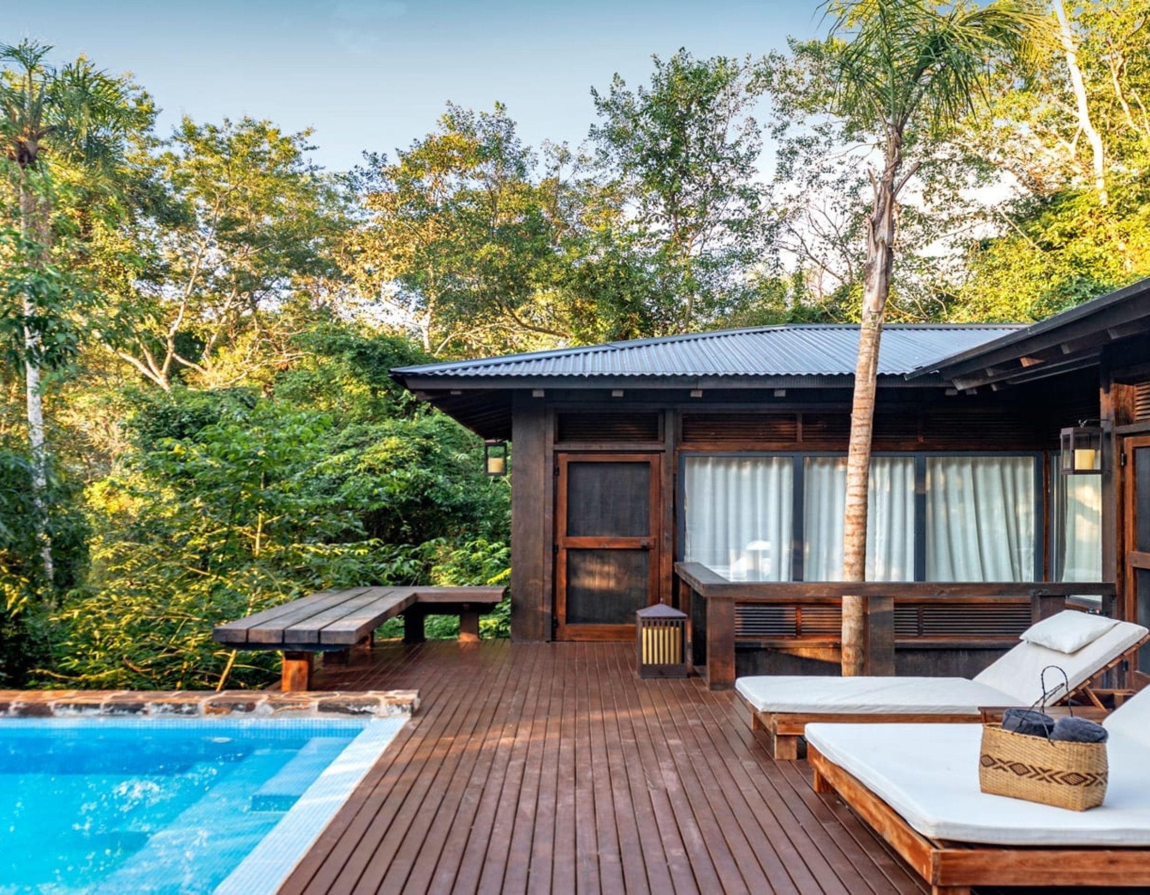 A modern wooden deck surrounds a small swimming pool, with a lounge chair and a table set nearby. Lush greenery and trees create a tropical backdrop. The house features large windows with sheer curtains, suggesting a serene outdoor living space.