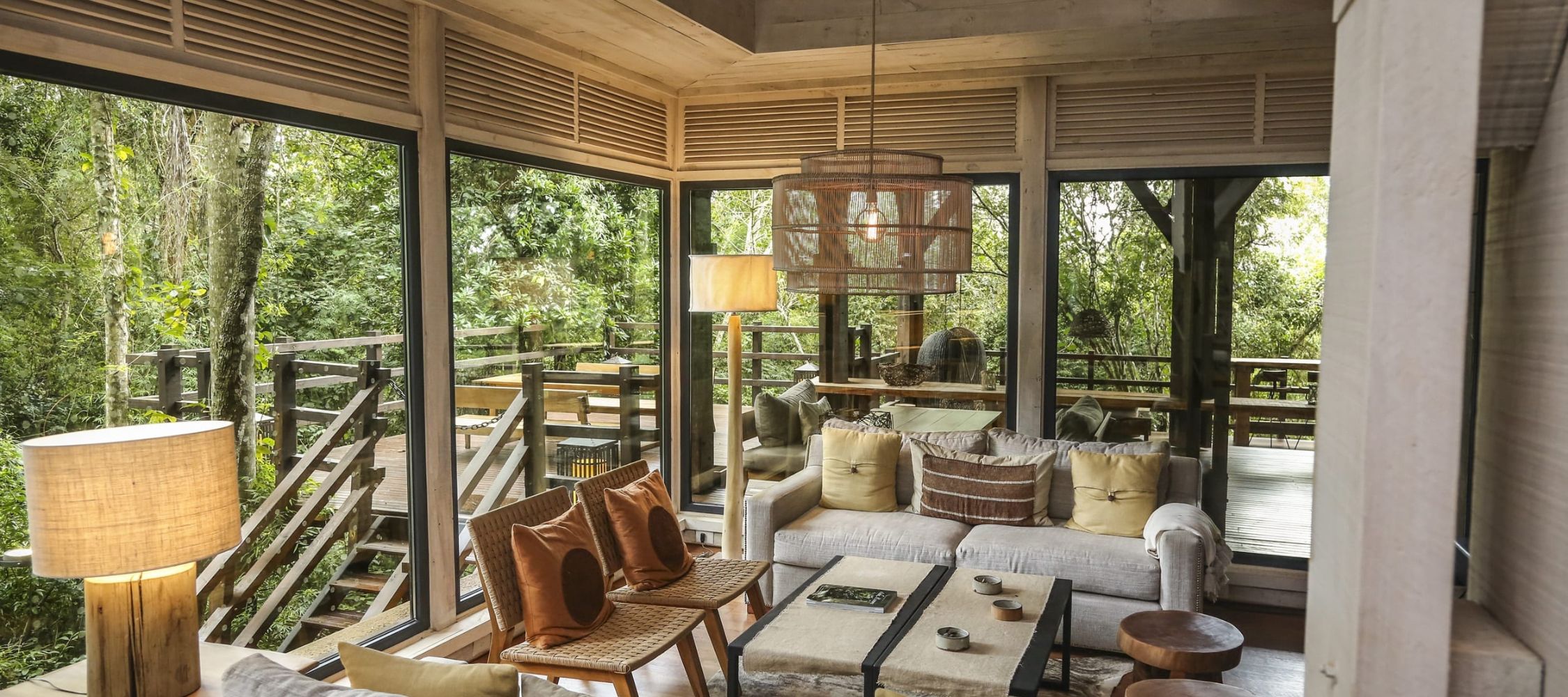 A cozy and modern living space with large windows overlooking a forest. The room features a seating area with plush sofas, a wooden coffee table, and stylish lighting. Natural elements and soft furnishings create a warm, inviting atmosphere.