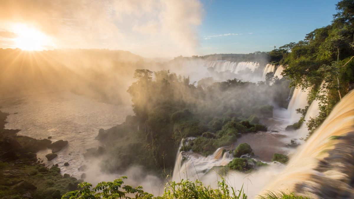 Majestic waterfalls cascading through lush greenery, with sunlight breaking through the mist and illuminating the scene. A serene river flows in the foreground, surrounded by vibrant vegetation and dramatic cliffs.