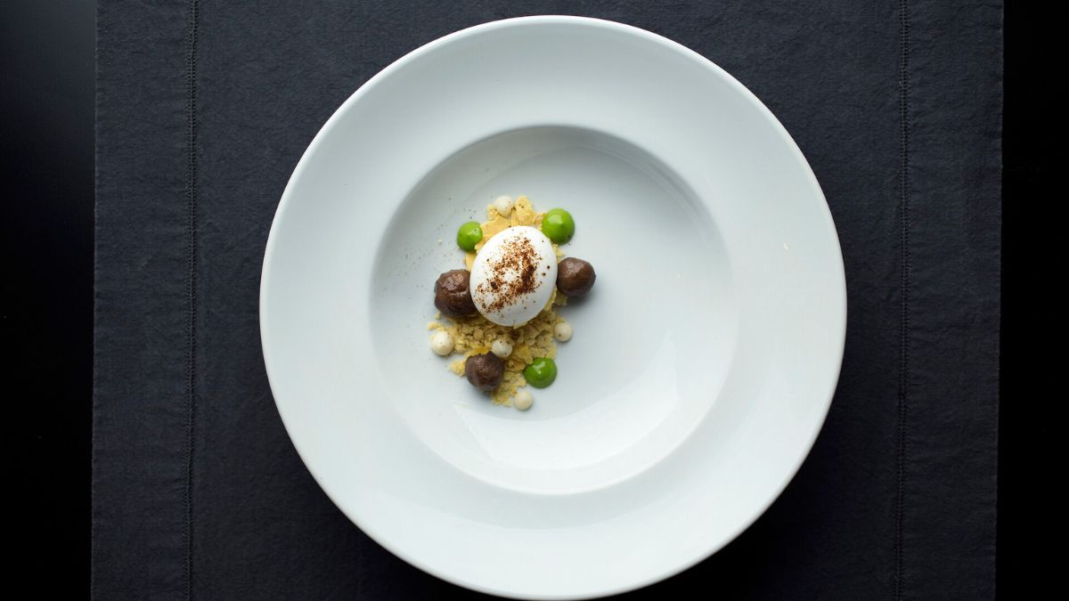 A gourmet dessert featuring a smooth white dome topped with chocolate shavings, surrounded by small dollops of chocolate mousse, crunchy bits, and green dots of sauce on a stark white plate.