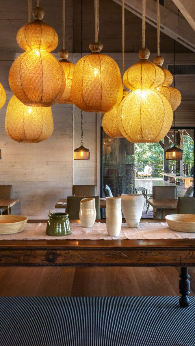A cozy dining space featuring large, warm-toned pendant lights hanging from the ceiling. A wooden table is adorned with decorative vases in various shapes and colors. The background shows comfortable seating and natural light filtering in through large windows.