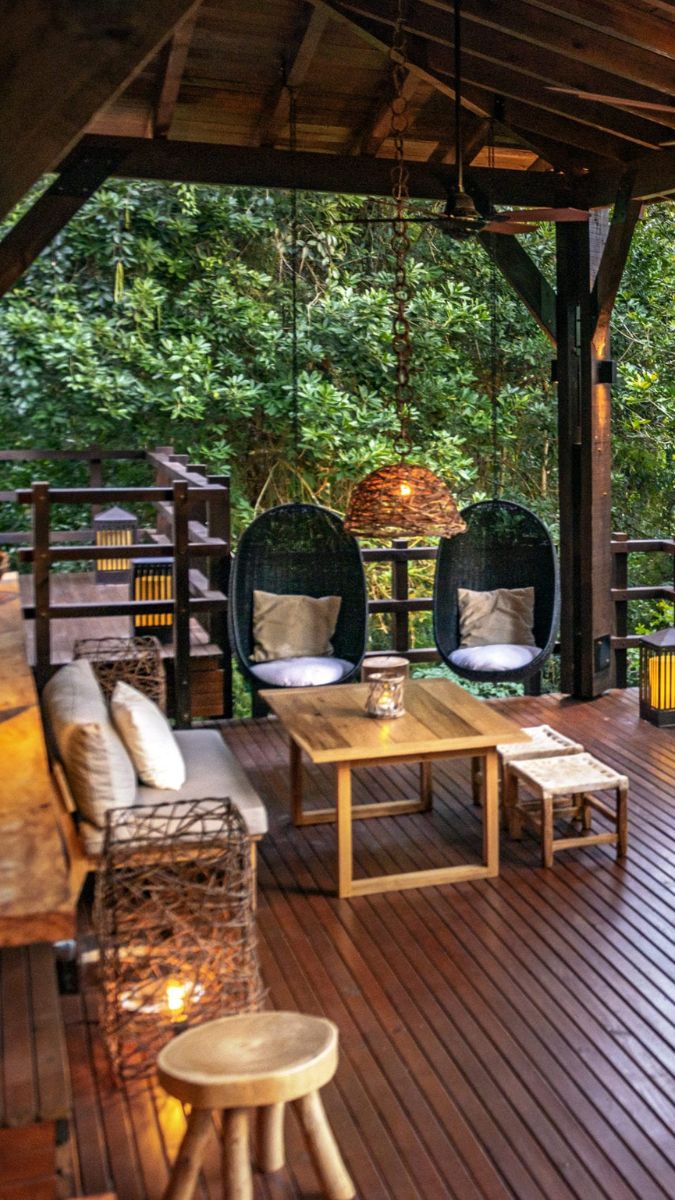 A cozy outdoor seating area featuring two hanging chairs, a wooden table, and soft lighting amidst lush greenery. The wooden deck is complemented by decorative lanterns and natural decor.