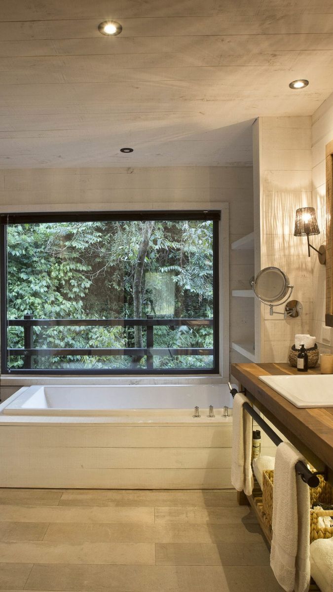 Modern bathroom featuring a large soaking tub and a wooden countertop with a sink. A large window offers a view of lush greenery outside. Soft lighting enhances the relaxing atmosphere.