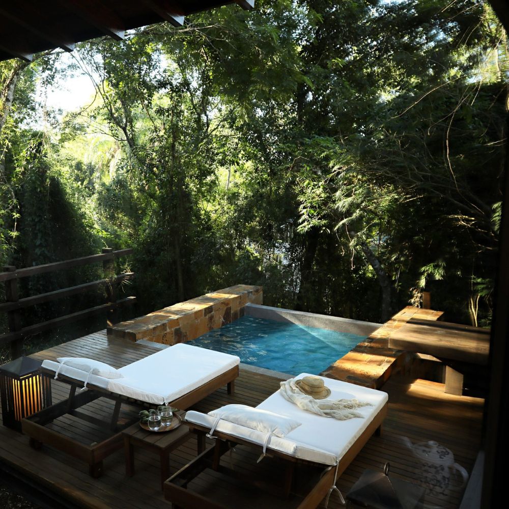 A serene outdoor area featuring two lounge chairs beside a small pool, surrounded by lush greenery and trees. Sunlight filters through the leaves, creating a tranquil atmosphere.