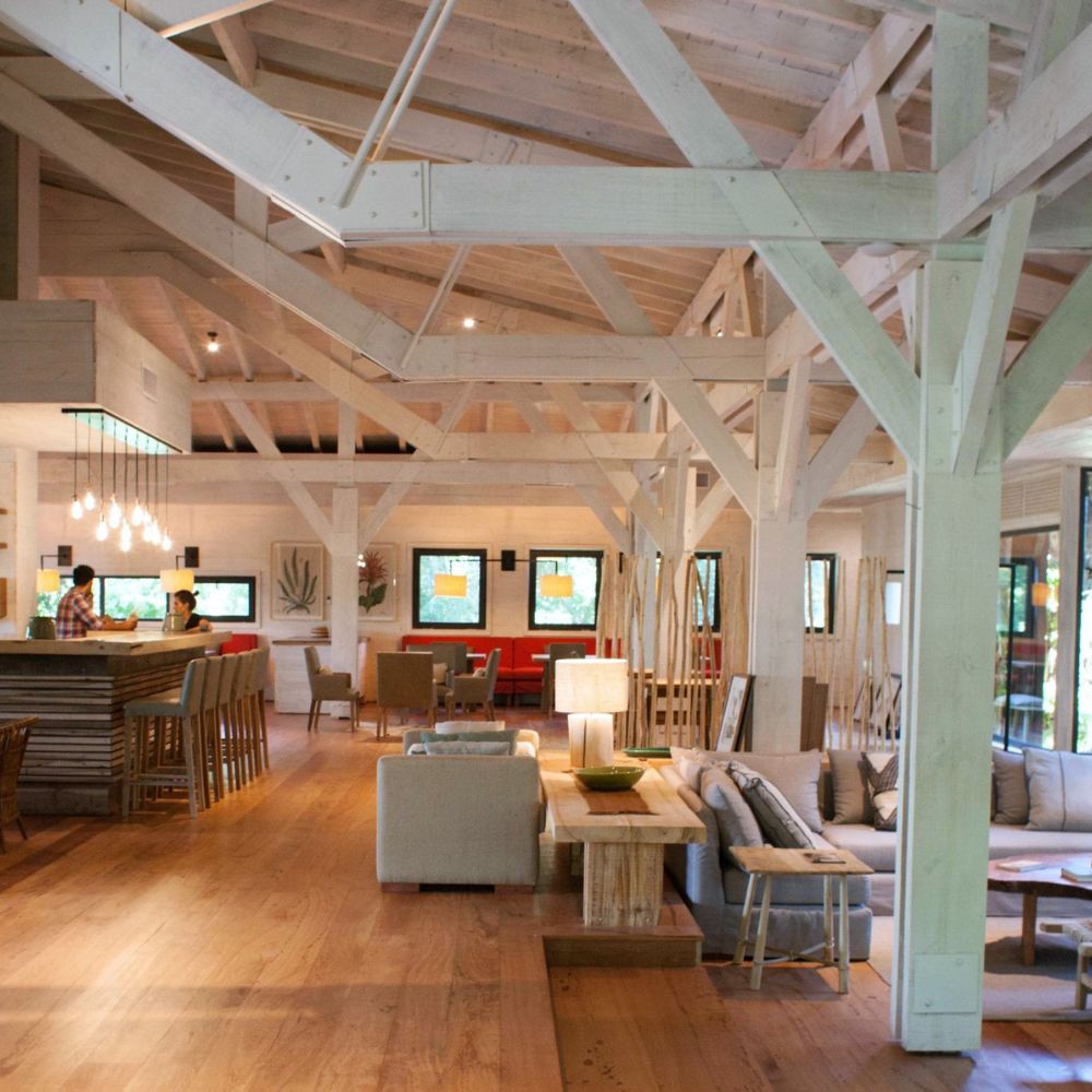 A spacious, modern interior with wooden beams, featuring a cozy seating area, a dining space with barstools, and large windows that allow natural light to fill the room.