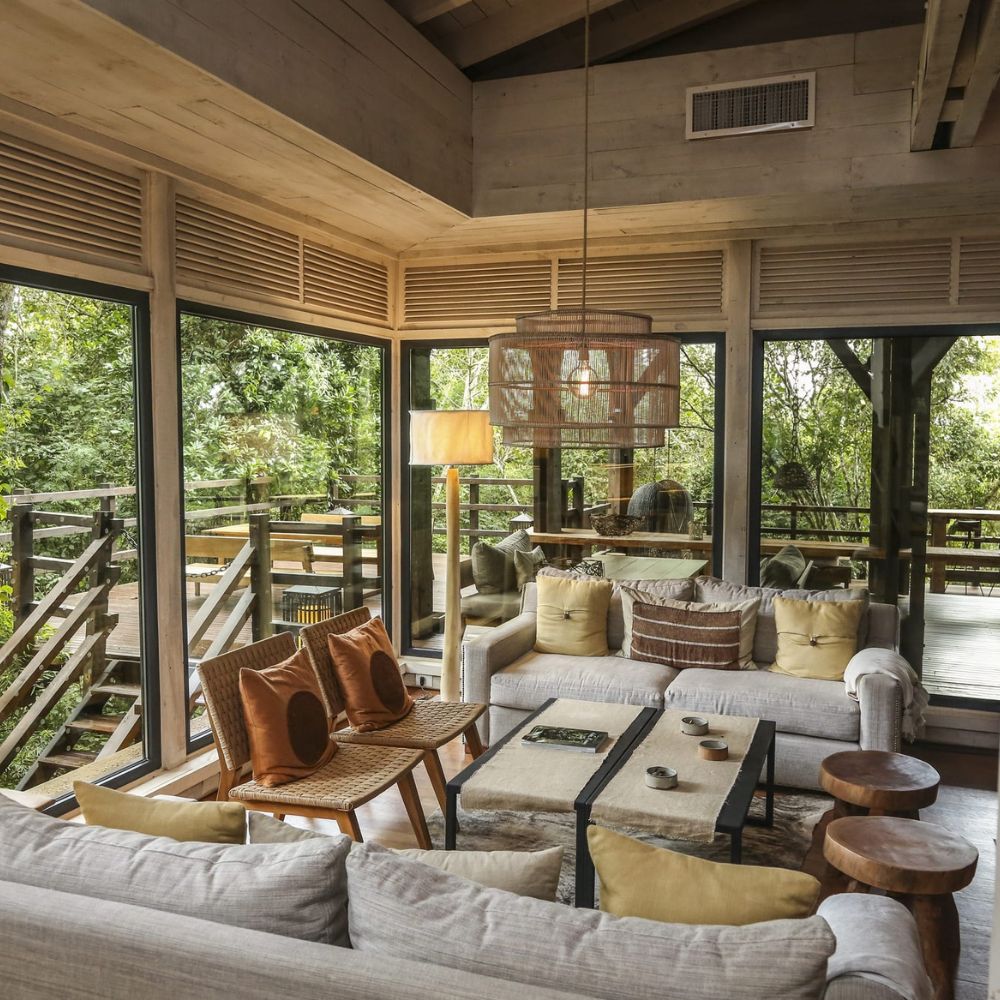A cozy living room featuring large windows that overlook a forested area. The space includes a light grey sofa with cushions, wooden side tables, and a stylish pendant light. Natural wood elements accentuate the modern design, creating a warm and inviting atmosphere.