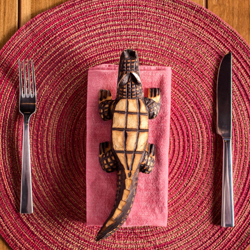 A wooden alligator sculpture rests atop a pink napkin, placed on a round, textured, pink placemat. A fork and knife are positioned on either side of the napkin.