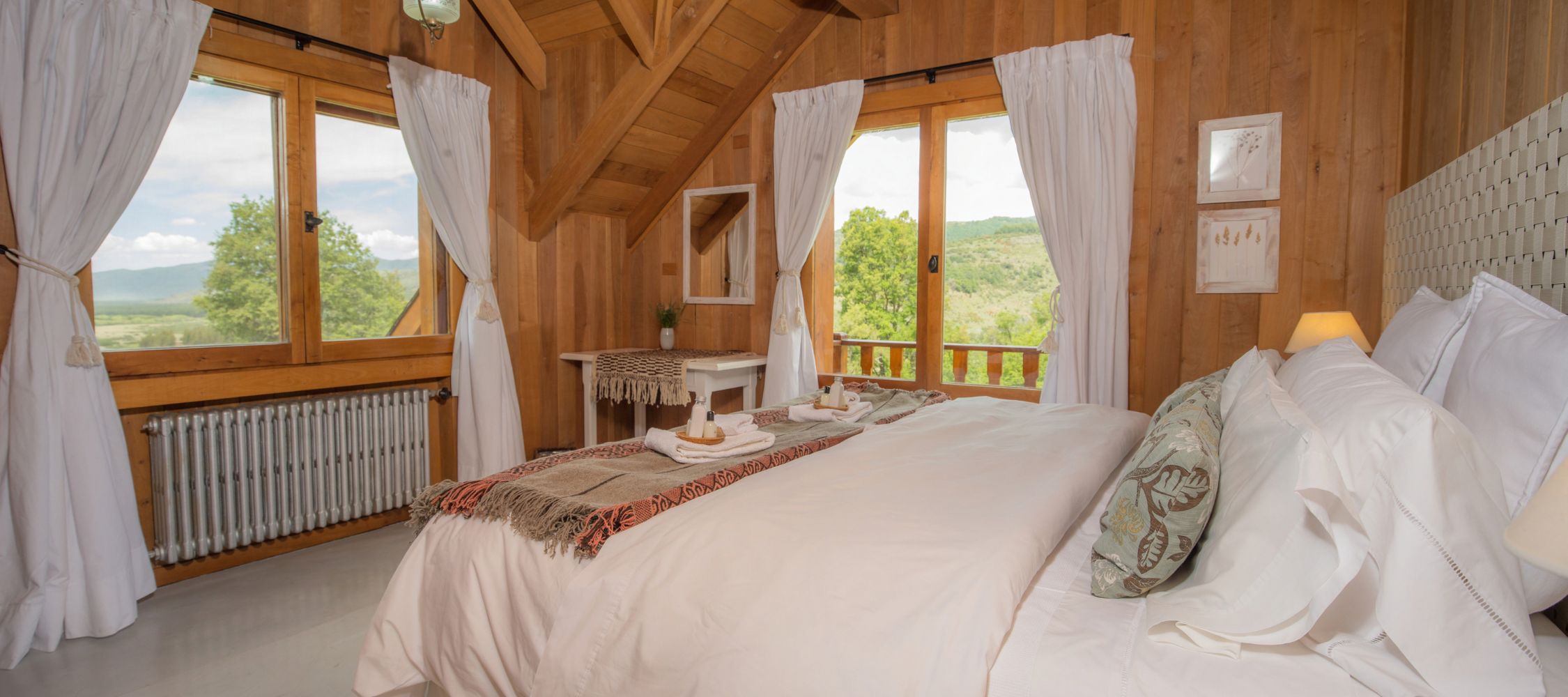 A cozy wooden bedroom featuring a large bed with white linens and decorative pillows. The room has a warm atmosphere with large windows offering a view of greenery and mountains. Soft lighting is provided by bedside lamps, and a small table is visible beside the bed. Elegant curtains frame the windows.