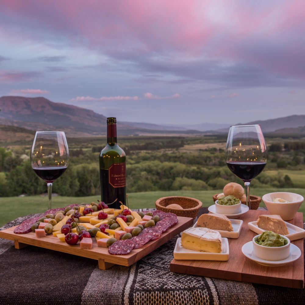 A wooden board filled with assorted cheeses, meats, olives, and grapes sits on a table with two wine glasses filled with red wine. A bottle of wine stands nearby, with a stunning landscape of rolling hills and a colorful sunset in the background.