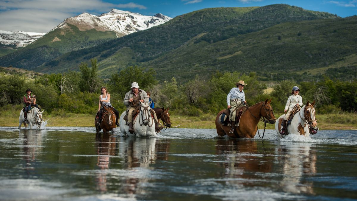 A group of five horseback riders crosses a shallow river, with snow-capped mountains and lush greenery in the background. The riders are dressed in casual outdoor attire, enjoying a sunny day in nature.