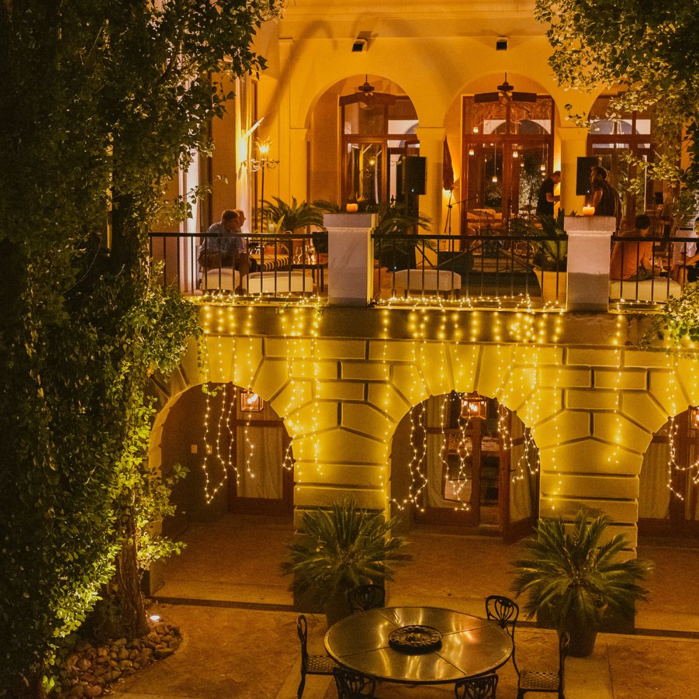 A beautifully lit courtyard adorned with string lights, featuring lush greenery and a round table with black chairs. The warm glow illuminates the surrounding arches and balconies, creating a cozy, inviting atmosphere.