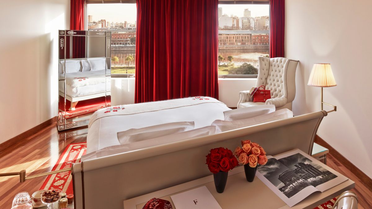 A luxurious hotel bedroom featuring a plush bed with white linens, a stylish armchair, a mirrored wall, and red curtains. A view of a city skyline is seen through large windows, and a decorative table holds fresh flowers and magazines.