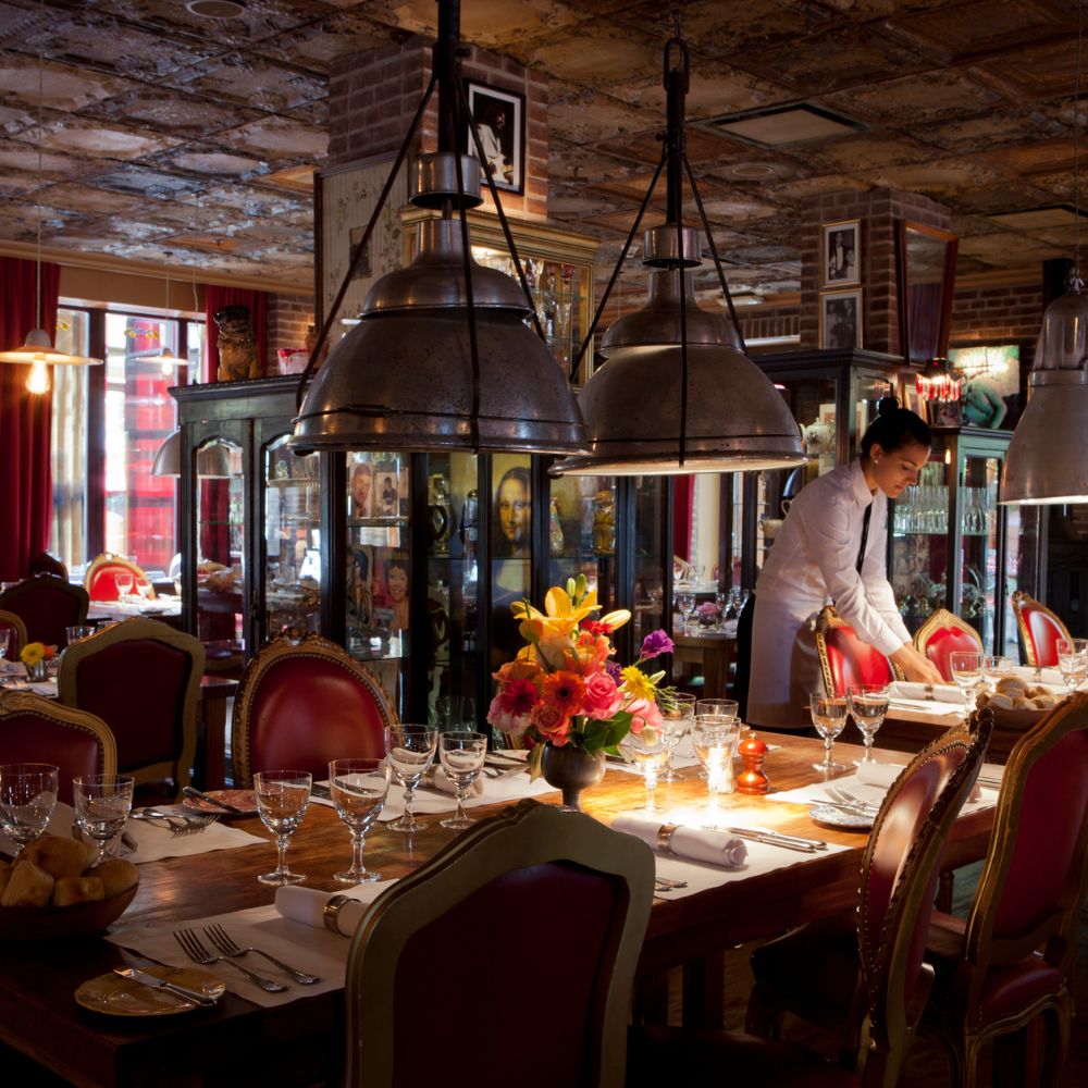 A warmly lit restaurant interior featuring a large wooden table set for dining, adorned with elegant glassware and a vibrant flower arrangement. A staff member is preparing the table, with vintage decor and glass display cases in the background, creating a cozy atmosphere.