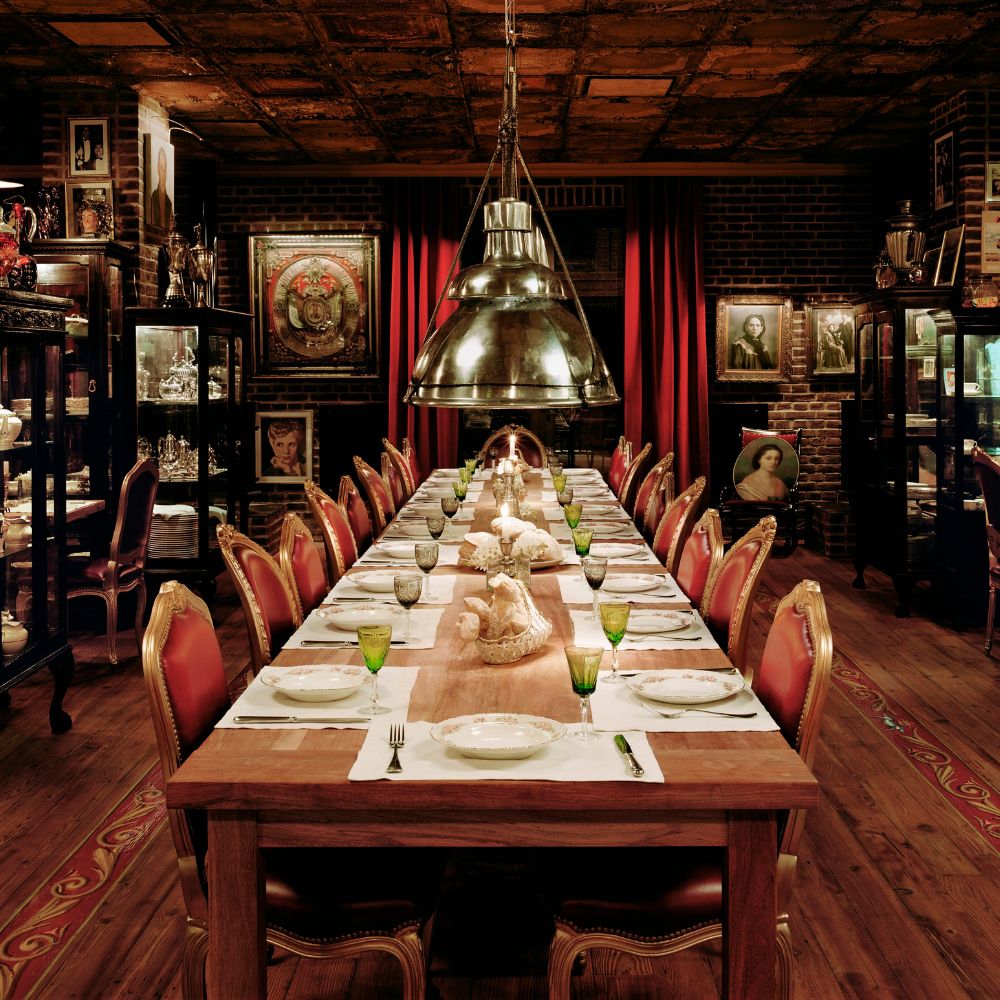 A long wooden dining table set with elegant green glassware and ornate tableware, surrounded by plush red chairs. The room features warm lighting, exposed brick walls, and showcases various antique artifacts and framed portraits in glass cabinets.
