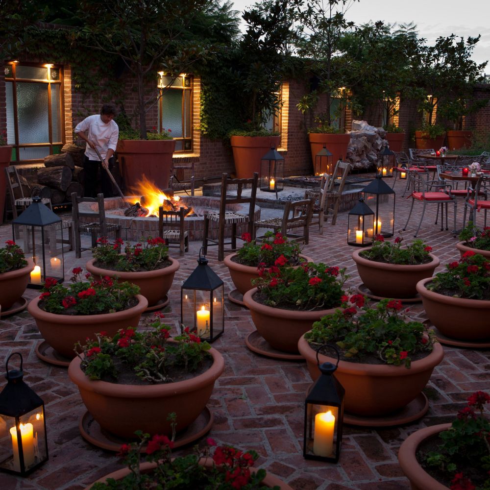 A tranquil outdoor courtyard with potted flowers and lanterns illuminating the space. A person is tending to a fire pit, surrounded by rustic chairs and tables. Soft lighting creates a warm, inviting atmosphere.