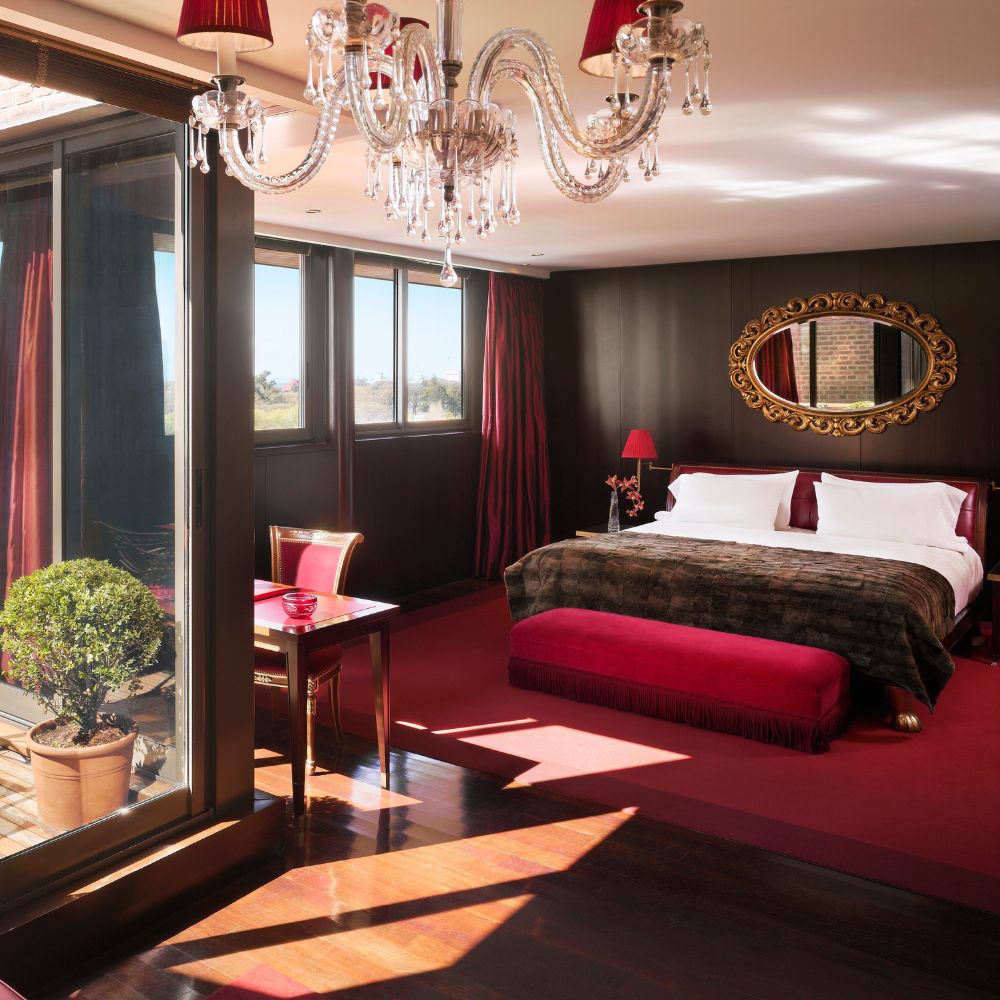 A luxurious hotel room featuring a large bed with a plush brown throw, accentuated by red decor. A decorative chandelier hangs above, and large windows provide natural light. A small dining table sits beside a potted tree, adding a touch of greenery to the elegant space.