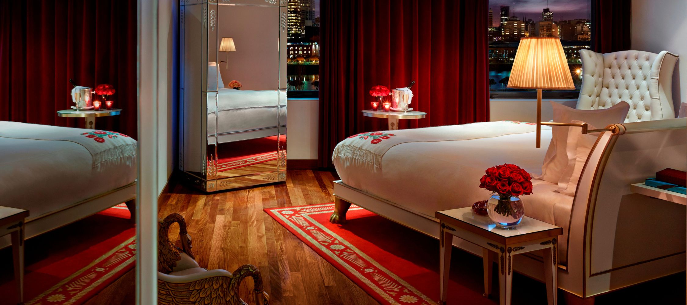 A stylish bedroom with a plush bed featuring a white blanket and decorative pillows. There is a large mirror reflecting the room, a vintage armchair, and a bedside table with a vase of red roses. The curtains are deep red, and the room has ambient lighting with city views through the window.