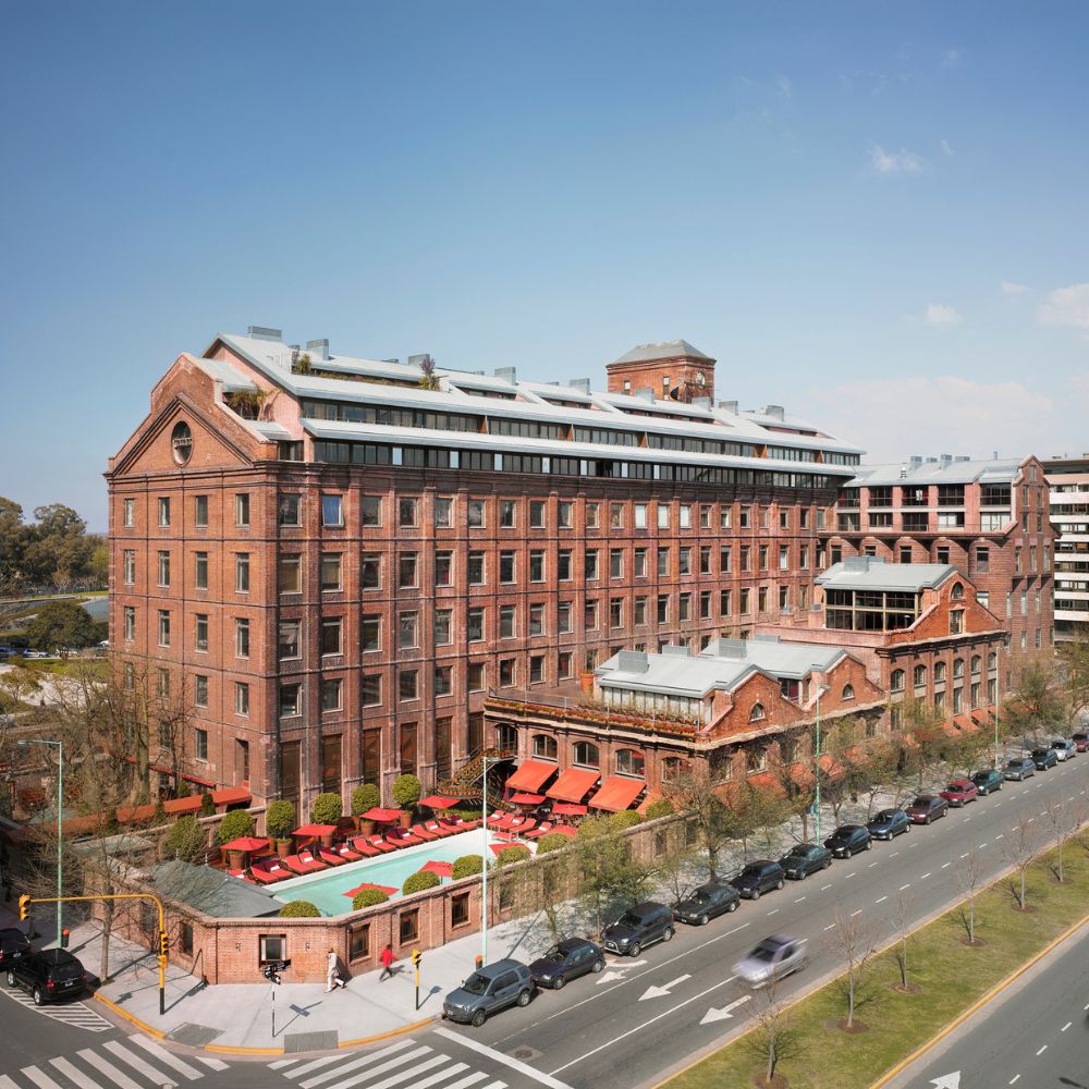 A historic red brick building with multiple stories, featuring large windows and a flat roof. The exterior is complemented by a landscaped area with red umbrellas on a patio. Modern vehicles are parked along the street in front of the building, and the sky is clear and blue.