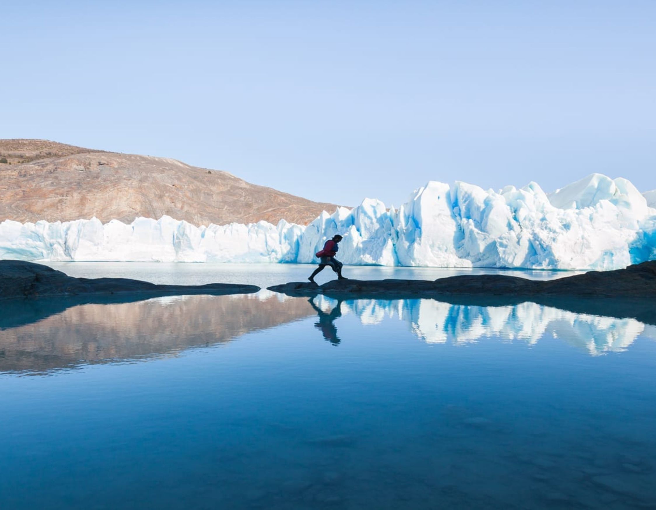 A person in a red jacket runs across a rocky outcrop near a calm, reflective body of water, with towering blue and white icebergs in the background against a clear sky.