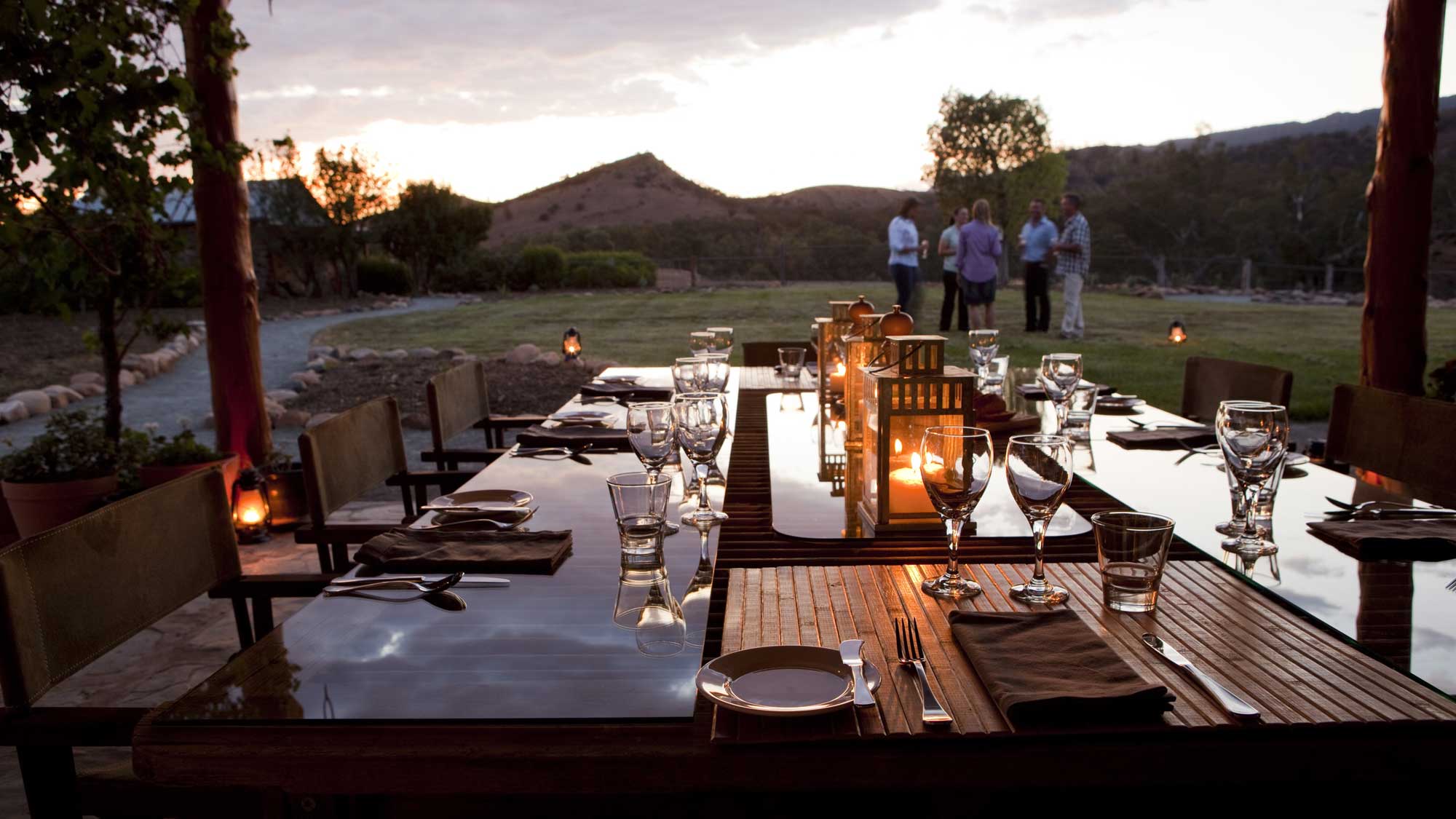 An elegantly set outdoor dining table with glassware and silverware, illuminated by soft candlelight at dusk. In the background, people converse near a scenic landscape of hills and trees.
