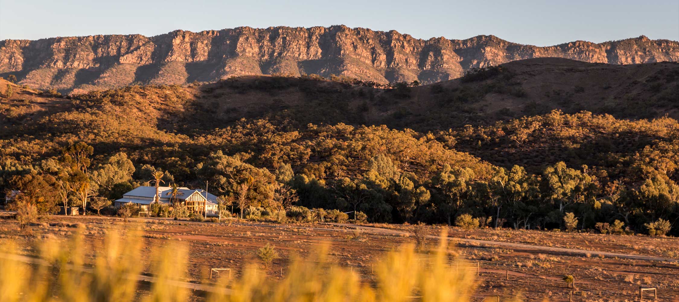 A rural landscape featuring a charming house nestled among lush trees, with rugged mountains rising dramatically in the background. The golden light of sunset casts a warm glow over the scene, highlighting the natural beauty.