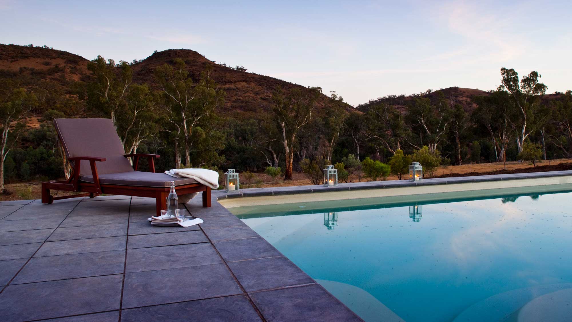 A tranquil poolside scene featuring a wooden lounge chair with a towel draped over it, next to a calm, reflective pool. Lanterns illuminate the area, with rolling hills and eucalyptus trees in the background during twilight.