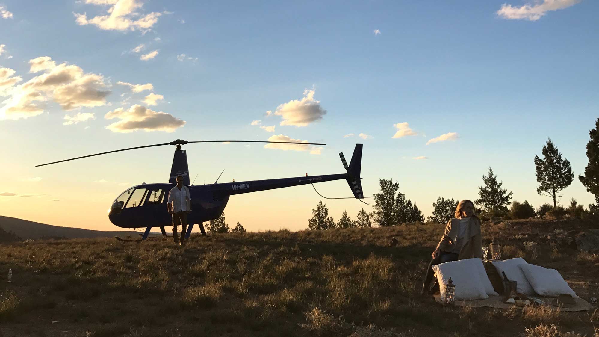 A blue helicopter on a grassy field during sunset, with a person standing next to it and another person sitting nearby on a cushion. Trees are visible in the background against a colorful sky.