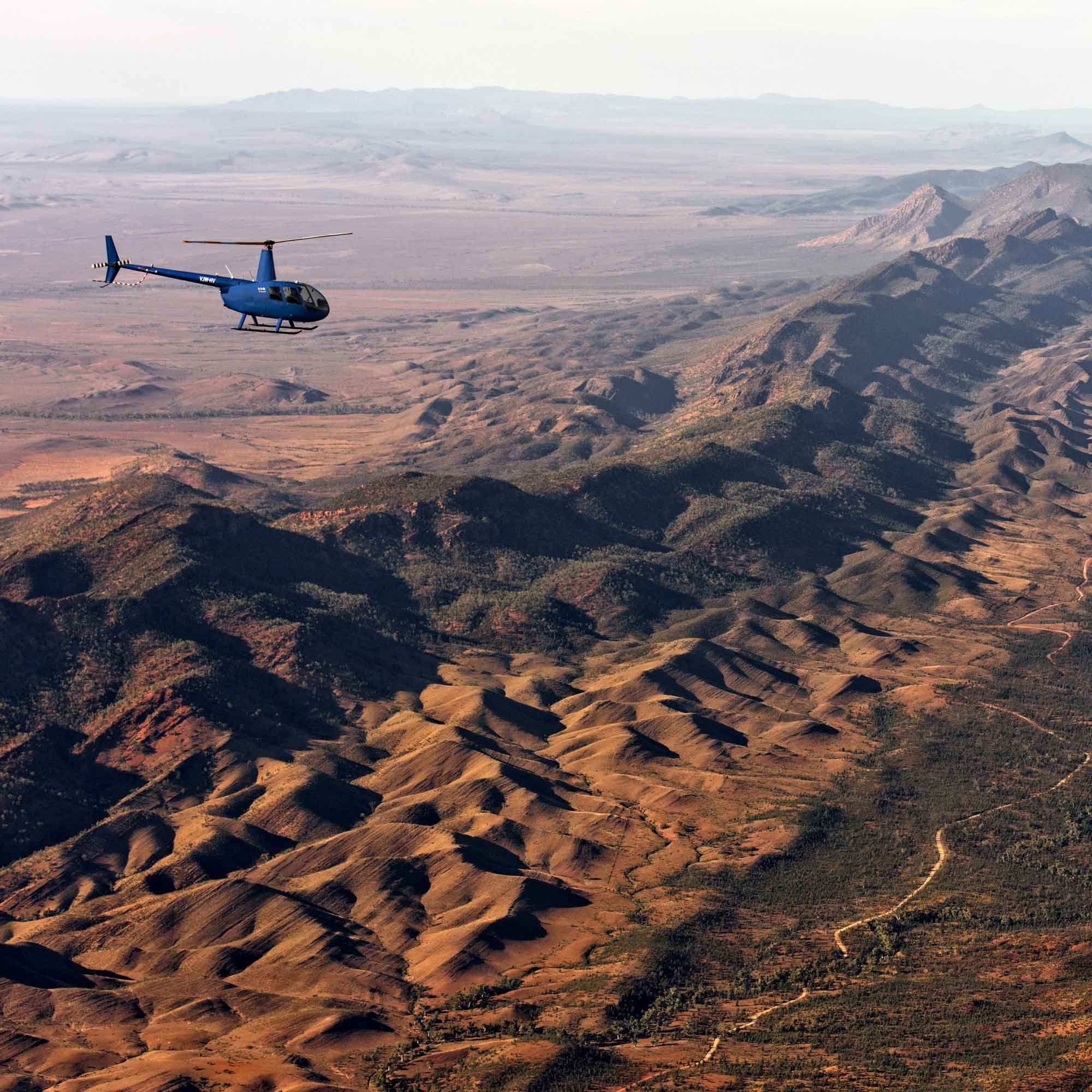 A blue helicopter flies over a vast, arid landscape with rolling hills and mountains in the background. The terrain features sandy slopes and sparse vegetation, stretching into the distance.