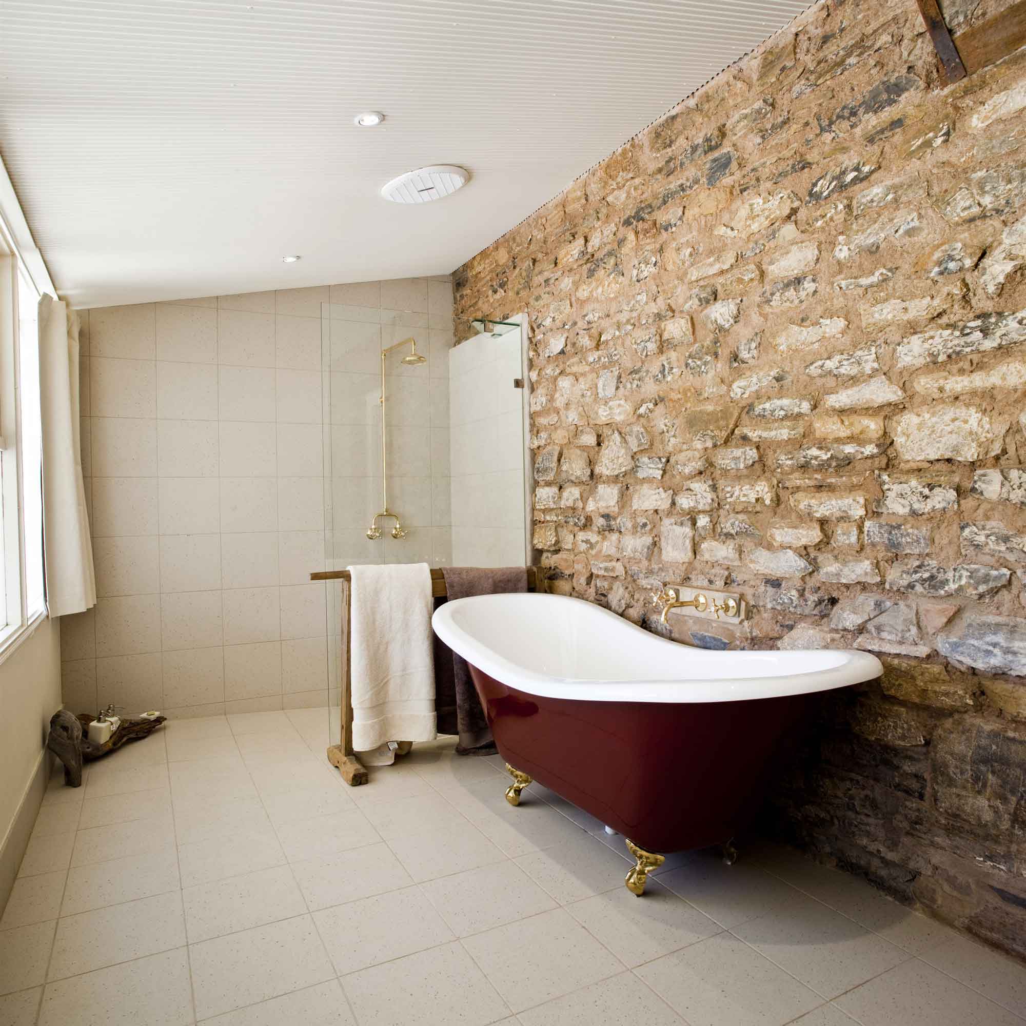 A modern bathroom featuring a freestanding red and white bathtub with gold fixtures. The room has a combination of textured stone walls and smooth tile flooring, complemented by natural light from a window. A glass shower enclosure is visible in the corner, and a towel rack hangs nearby.