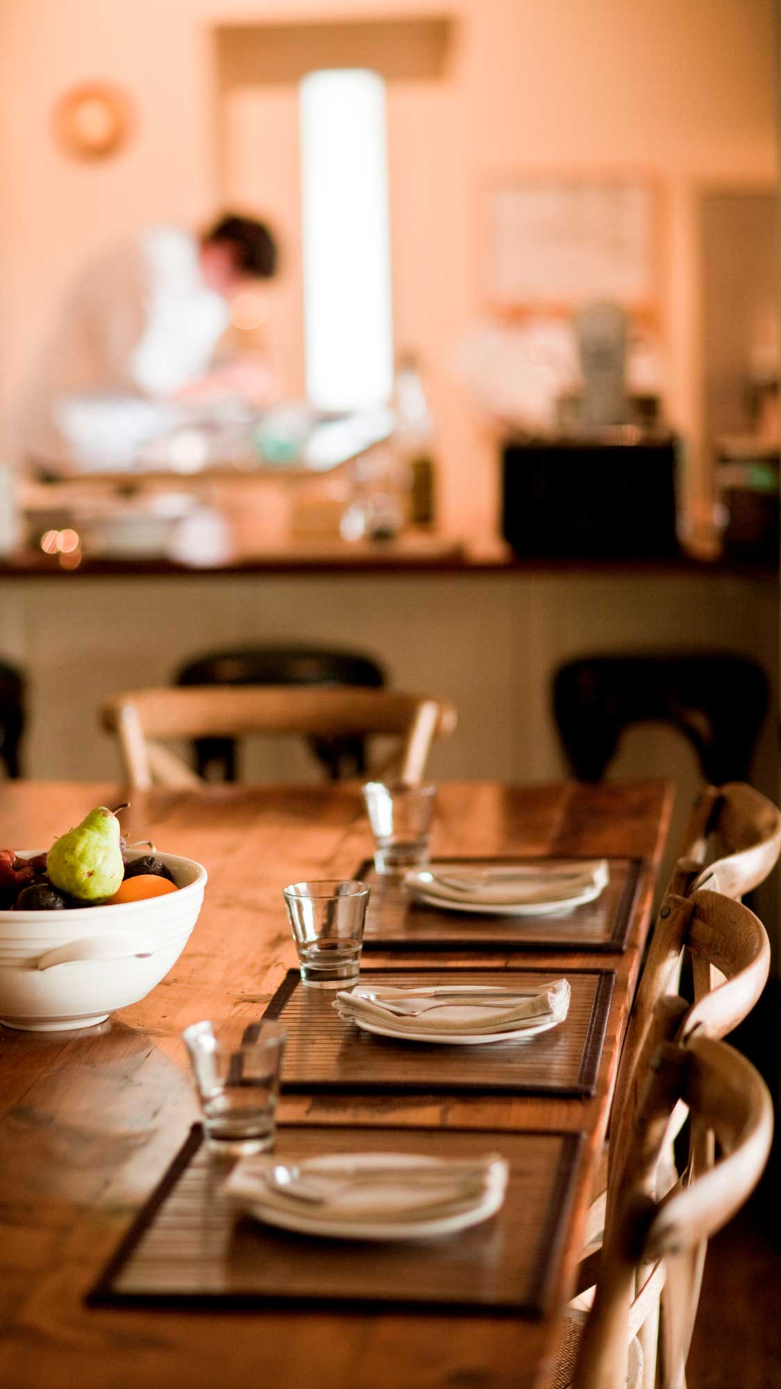 A wooden dining table set with three white plates, glasses, and a bowl of fruit in the foreground. Soft lighting creates a warm atmosphere. A person can be seen preparing food in the background.