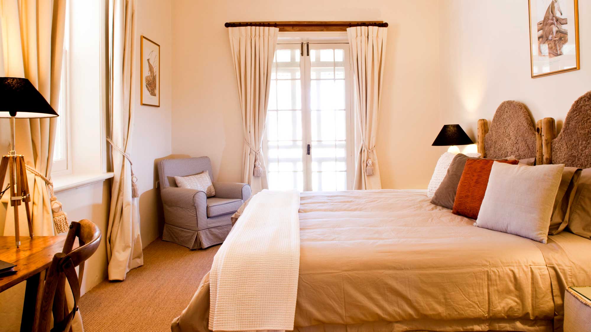 A well-decorated bedroom featuring a large bed with soft bedding, two bedside lamps, and a cozy armchair in the corner. Natural light fills the room through a set of double doors leading to a balcony. Walls are adorned with framed artwork, and a rustic wooden table is positioned nearby.