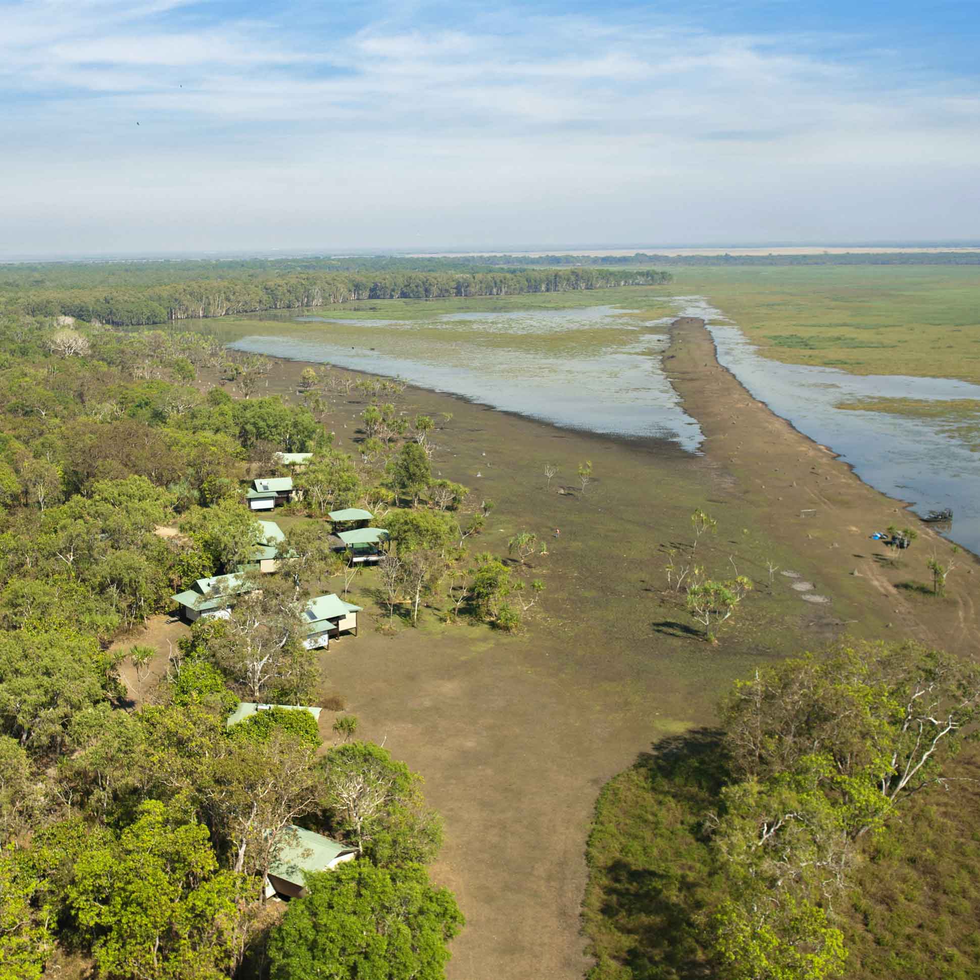 Aerial view of a lush green landscape with several small buildings nestled among trees, alongside a wide river and marshland. The sky is clear with a few clouds, showcasing the natural beauty of the area.