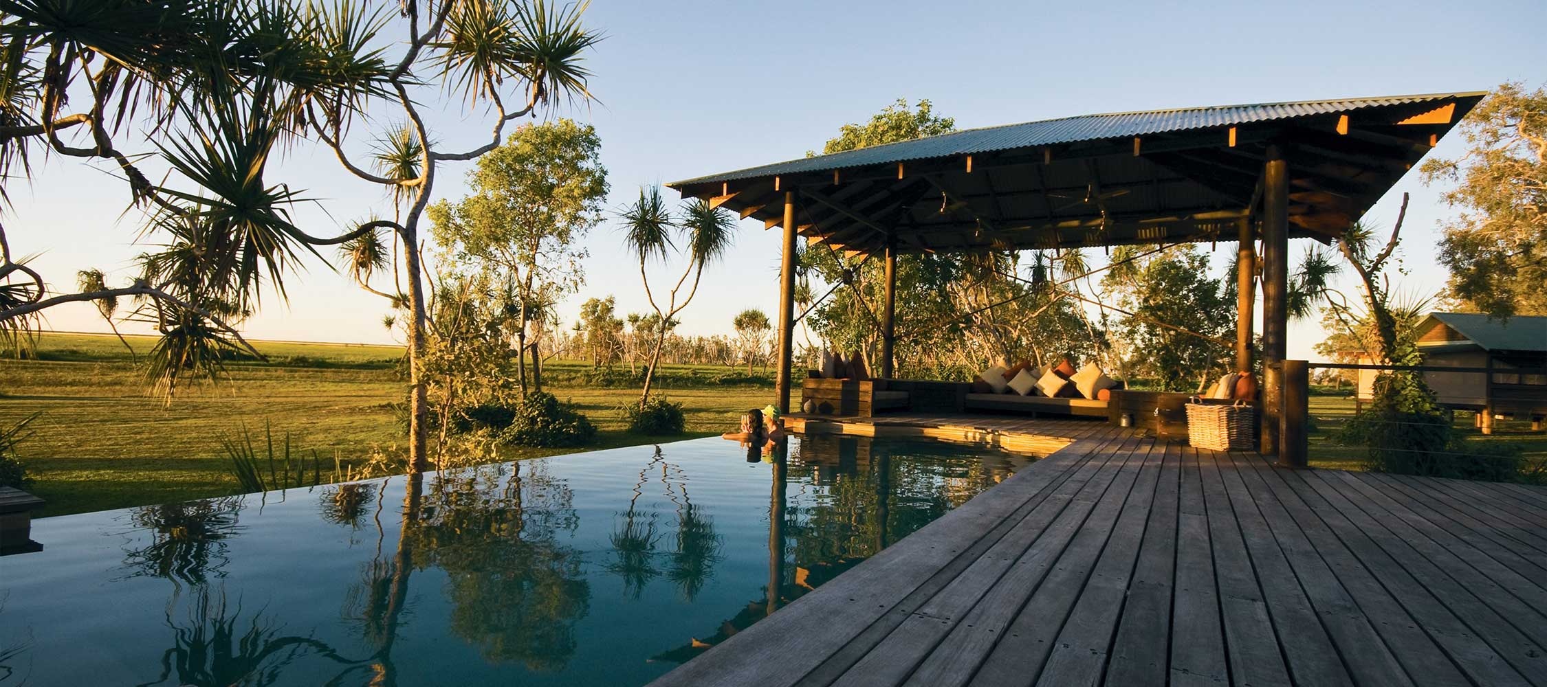 A serene outdoor setting featuring a wooden deck extending over a reflective pool. A gazebo with a sturdy roof offers a shaded seating area surrounded by lush greenery and palm trees. The horizon shows open fields under a clear sky.
