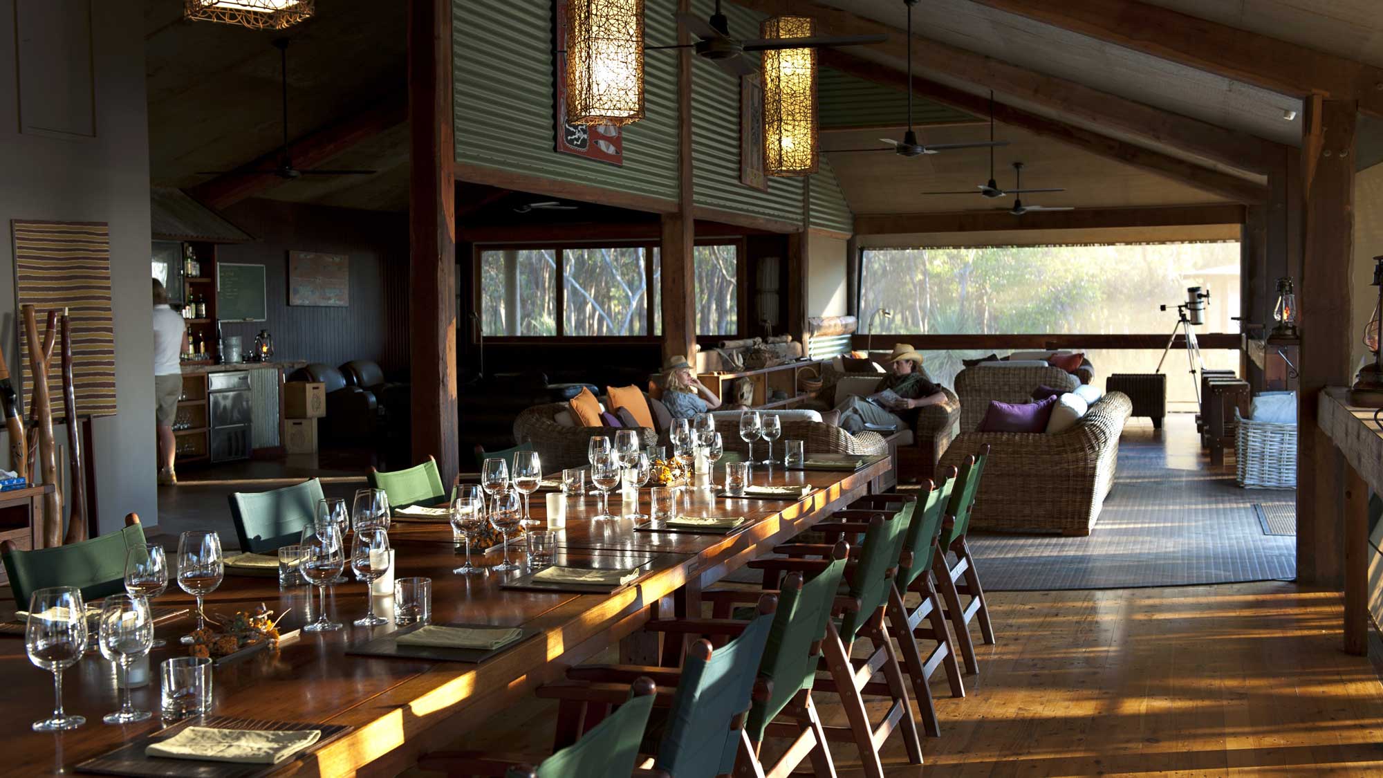 A spacious dining area with a long wooden table set with glasses and menus. Cozy seating areas with cushions are visible in the background, surrounded by large windows letting in natural light. Warm wooden beams and hanging lights add to the inviting atmosphere.