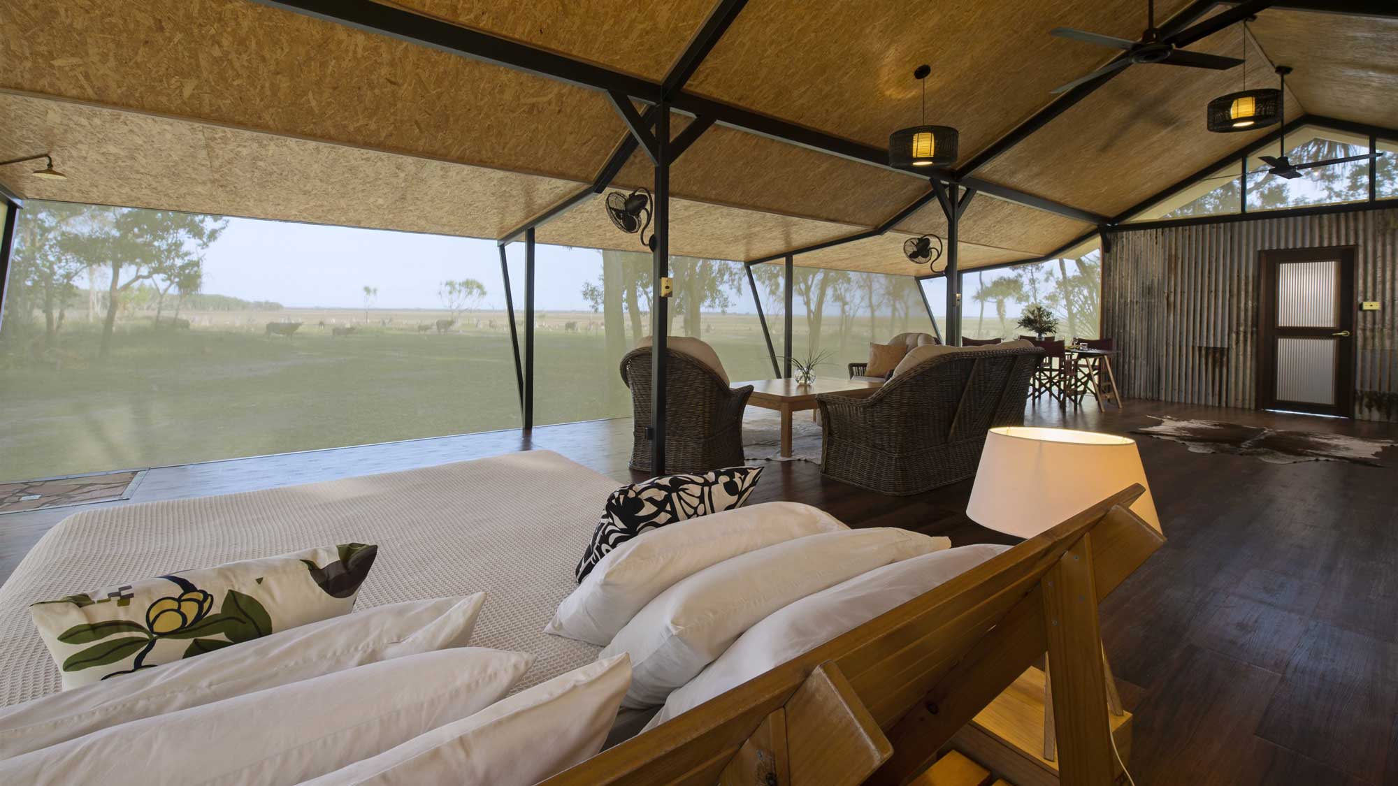 A cozy interior of a glamping tent featuring a large bed with decorative pillows, comfortable seating area with woven chairs, and wooden accents. Large windows allow views of a green landscape outside. A soft glow from a side lamp adds warmth to the space.