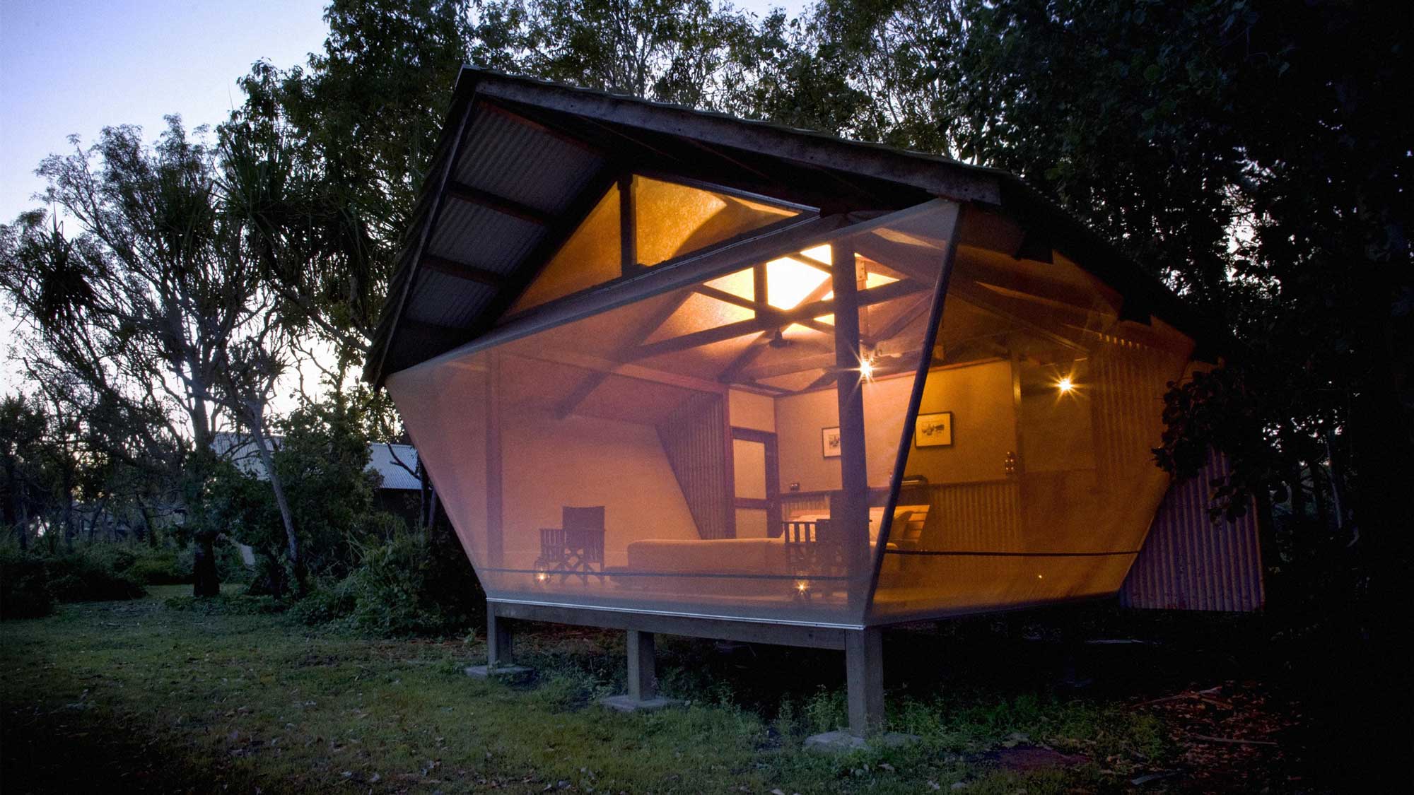 A modern, angular glass house sits among trees at dusk, showcasing warm interior lights. The unique structure features wooden beams and a raised foundation, blending natural elements with contemporary design.