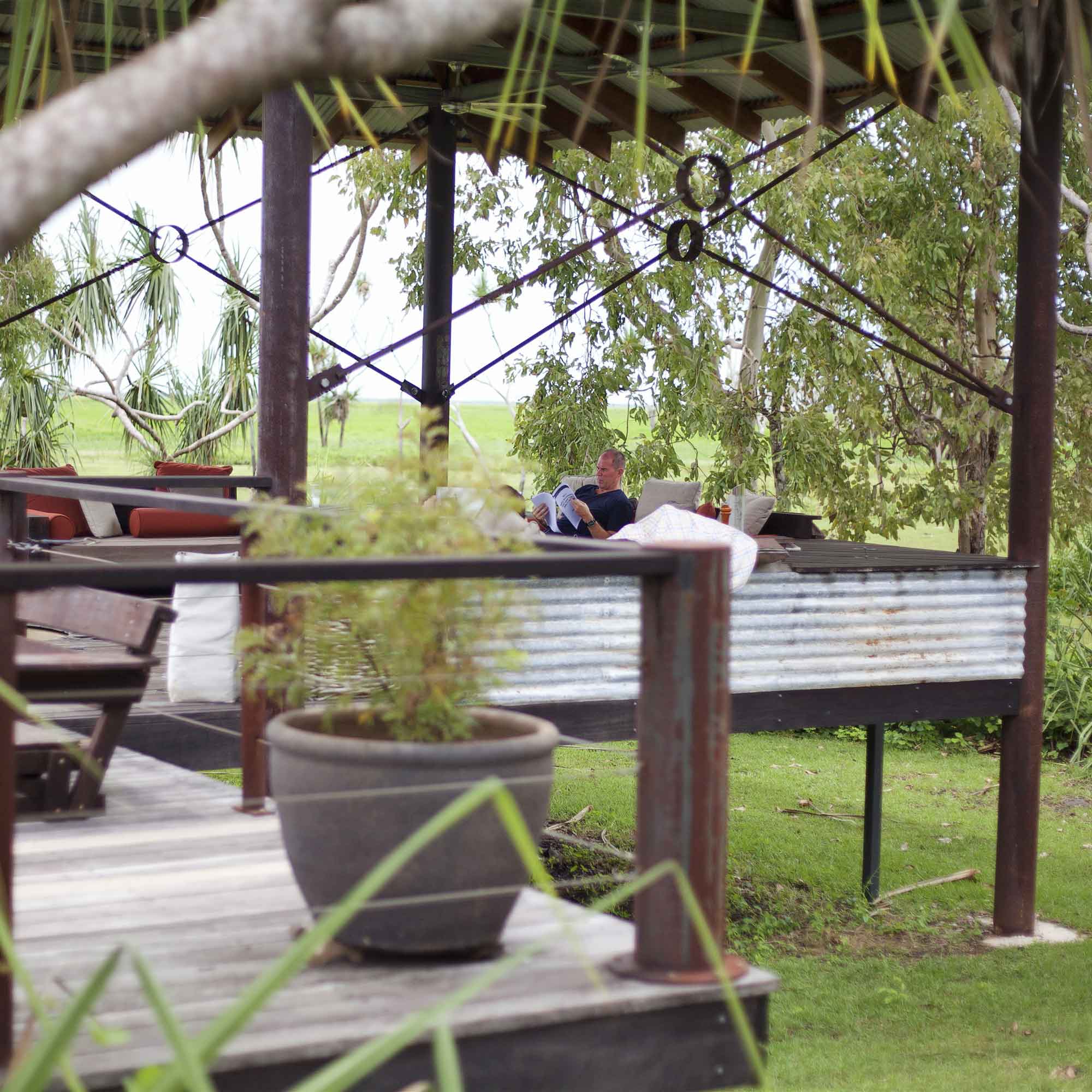 A person sits on a shaded terrace surrounded by greenery, relaxing on a couch with a blanket. The structure features wooden beams and corrugated metal accents.