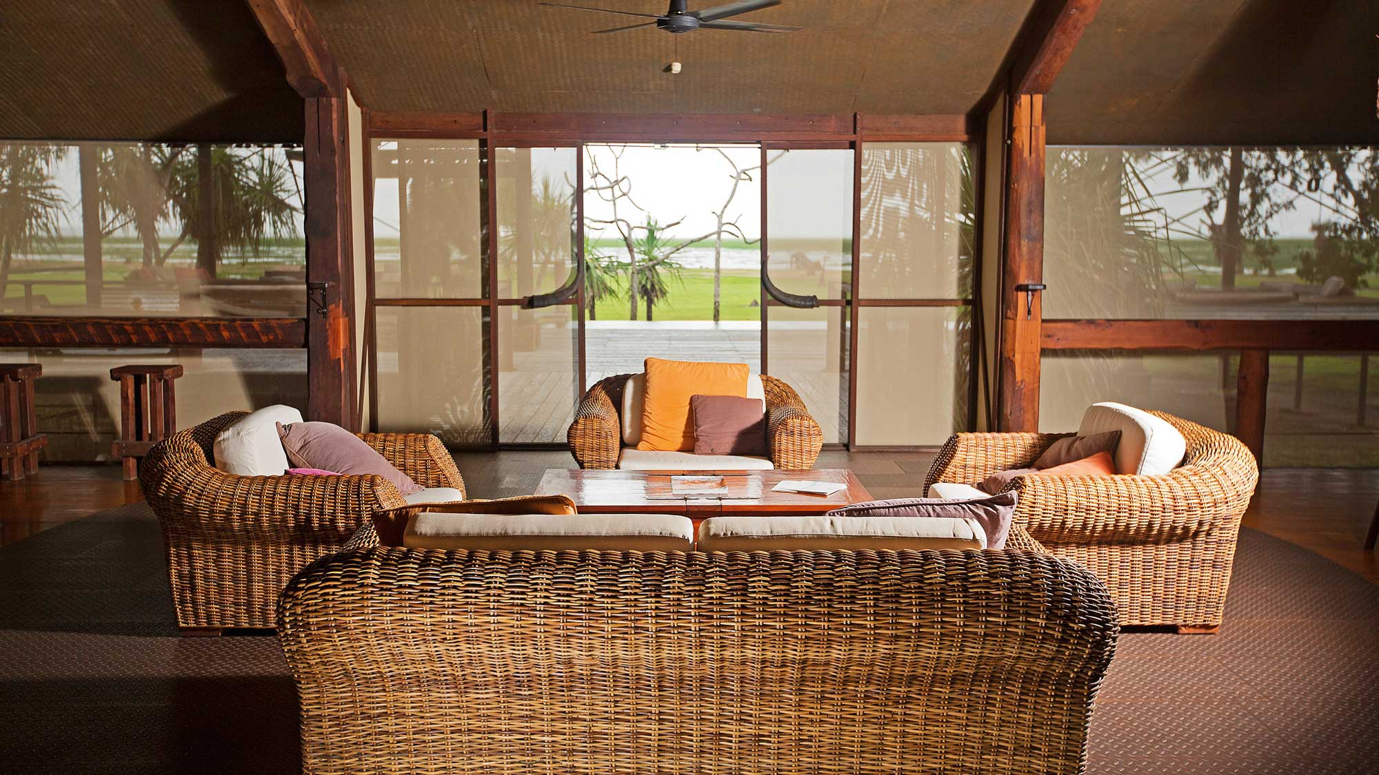 A cozy indoor seating area with woven rattan furniture, including a circular coffee table, surrounded by plush cushions. Large windows showcase a view of greenery and the ocean in the background.