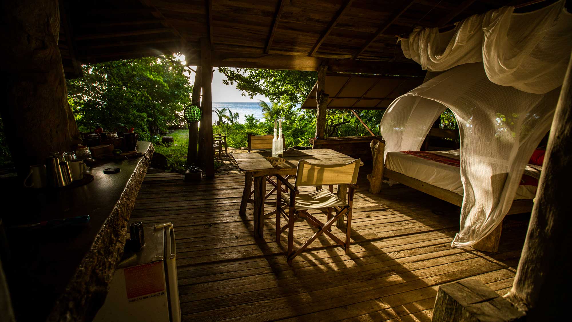 Cozy wooden cabin interior featuring a bed with a mosquito net, a dining table, and a scenic view of the ocean through open windows. Natural light floods the space, highlighting the rustic decor and lush greenery outside.