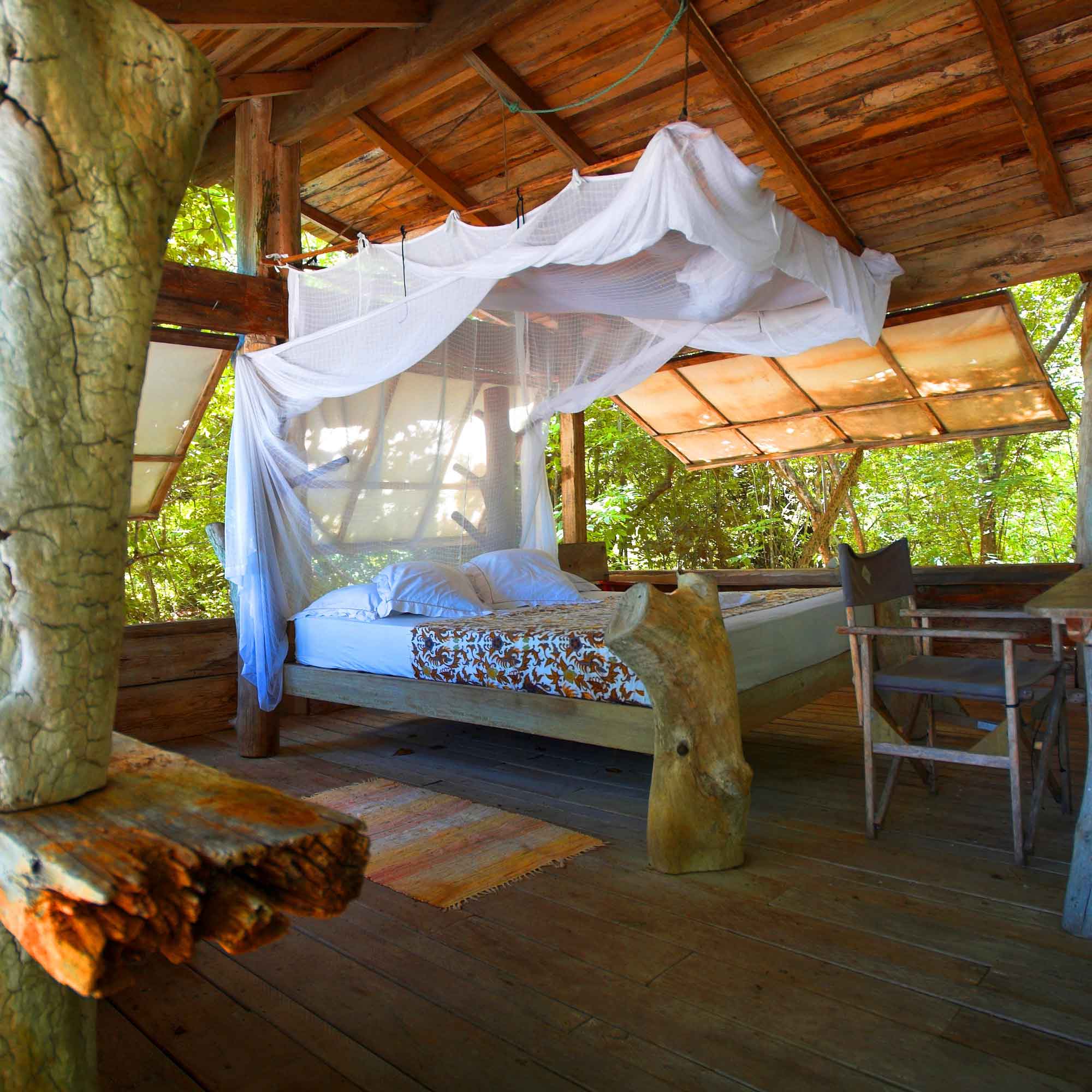 A cozy wooden bedroom featuring a bed draped with a white canopy. Natural light streams in through large windows and the space is surrounded by greenery. Rustic wooden furniture complements the serene atmosphere.