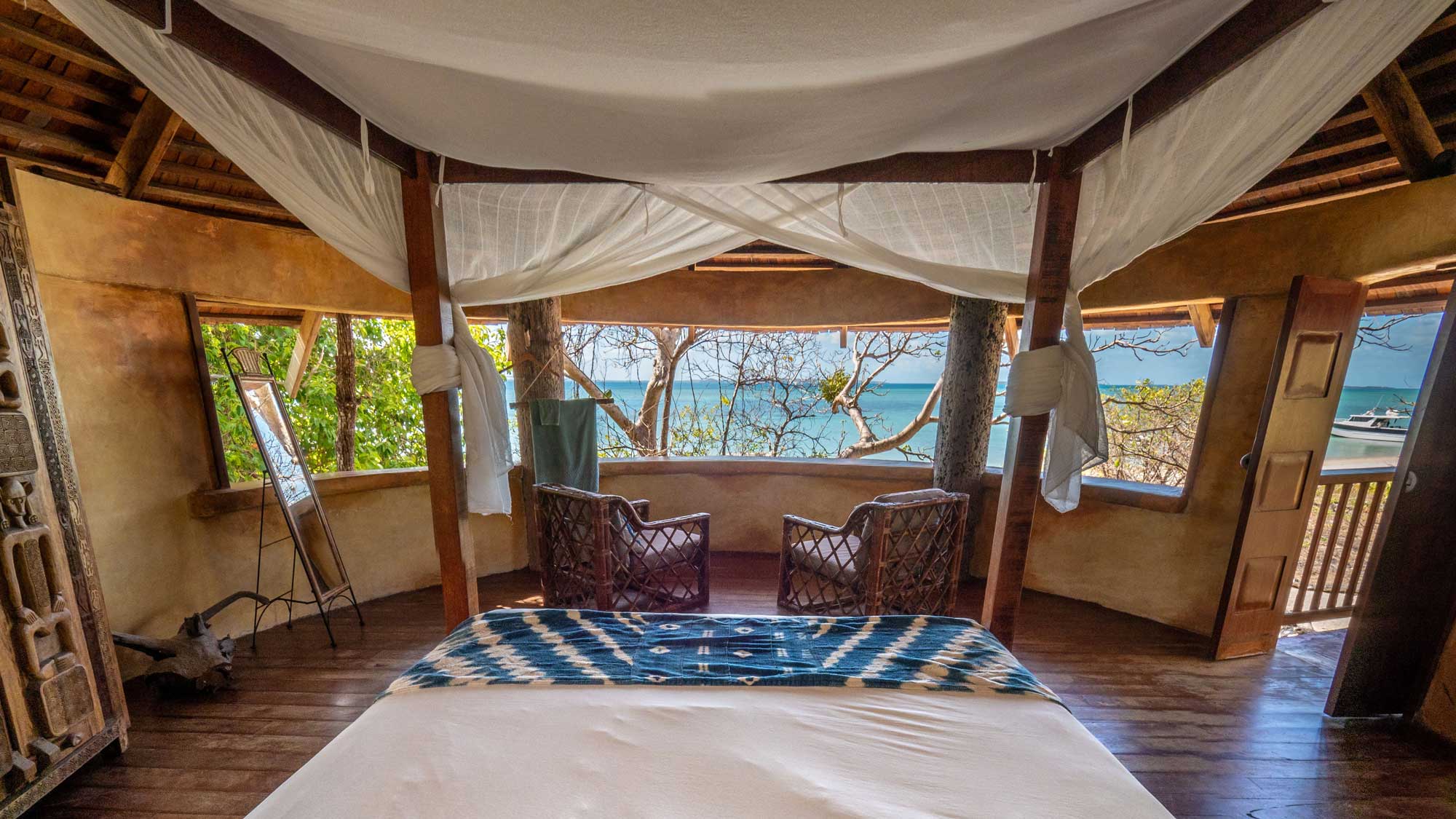 A serene bedroom with a wooden bed featuring patterned bedding. Two wicker chairs face large windows overlooking a tranquil sea and lush greenery. Natural light streams in, enhancing the warm, rustic decor.