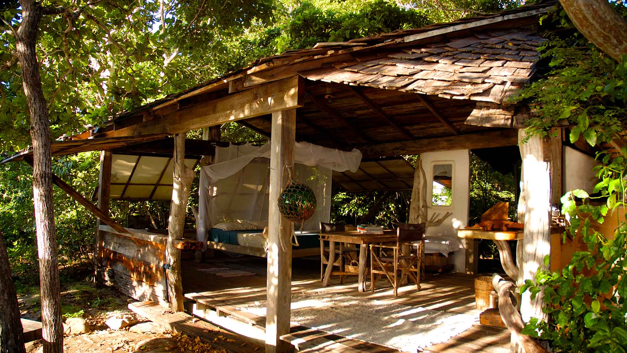 A rustic wooden cabin surrounded by greenery, featuring an open porch with a dining table and chairs, and a cozy bed draped with sheer curtains.